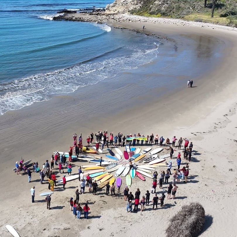A group gathers in a circle on the sand, with their boards inside the circle, before a memorial paddle out.