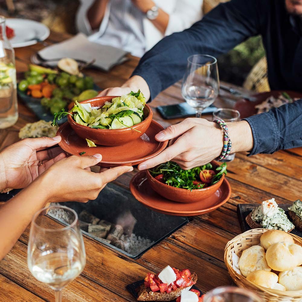 Image of a wooden table full of food and wine glasses. Two sets of hands are passing a dish of vegetables across the table. 